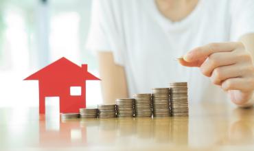 Small red house with stacks of coins next to it on a counter with a person behind it